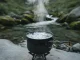 Kleiner schwarzer Campingtopf auf einem Gaskocher auf einem Felsen am Bach; das Wasser darin kocht sprudelnd, Dampf steigt senkrecht auf.