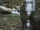 Weißer Hohlfaser-Wasserfilter an einem durchsichtigen Trinkbeutel, an einen Felsen am Bach gelehnt; gefiltertes Wasser läuft in einem dünnen Strahl in eine klare Flasche.