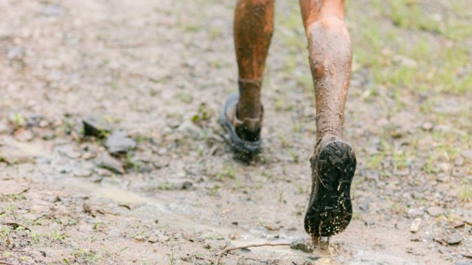 Nahaufnahme von hinten: schlammbedeckte Beine und Laufschuhe auf einem nassen, matschigen Waldweg, Wasser spritzt aus einer Pfütze