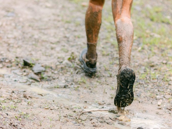 Nahaufnahme von hinten: schlammbedeckte Beine und Laufschuhe auf einem nassen, matschigen Waldweg, Wasser spritzt aus einer Pfütze