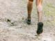 Nahaufnahme von hinten: schlammbedeckte Beine und Laufschuhe auf einem nassen, matschigen Waldweg, Wasser spritzt aus einer Pfütze